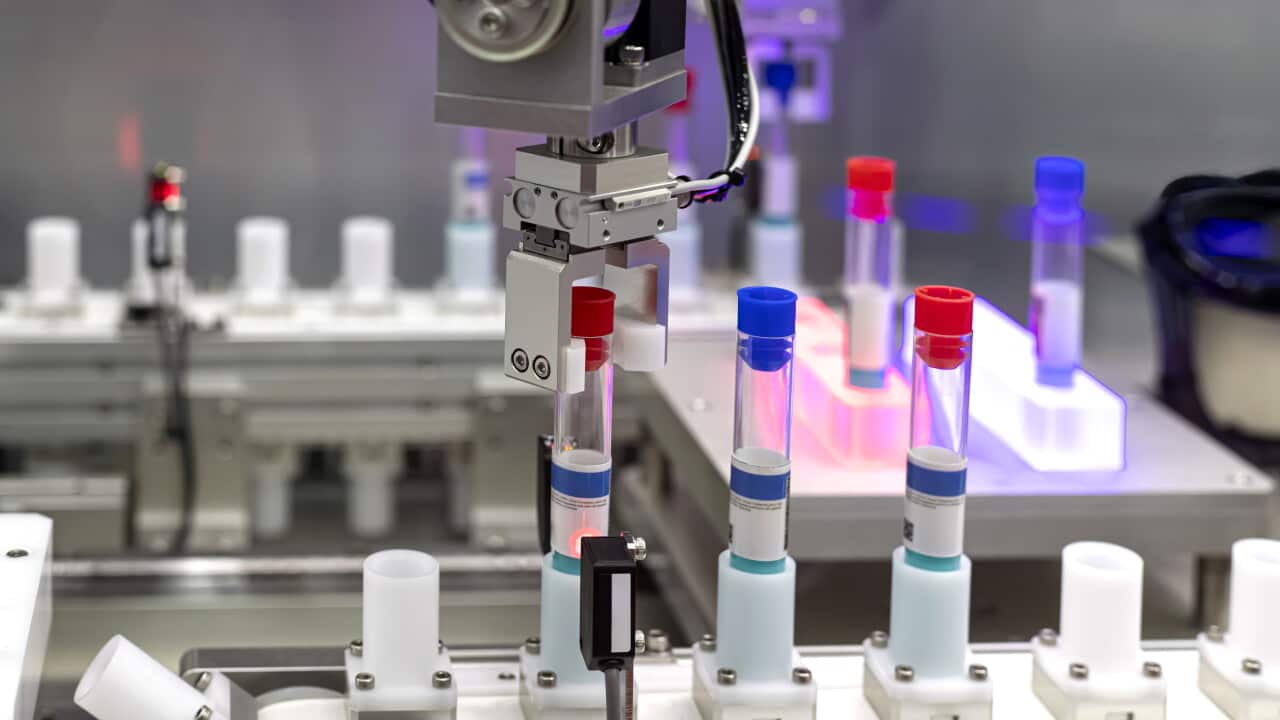A robotic arm gripper carefully positions a labelled, red-capped test tube into an automated conveyor slot within a high-tech laboratory setting.