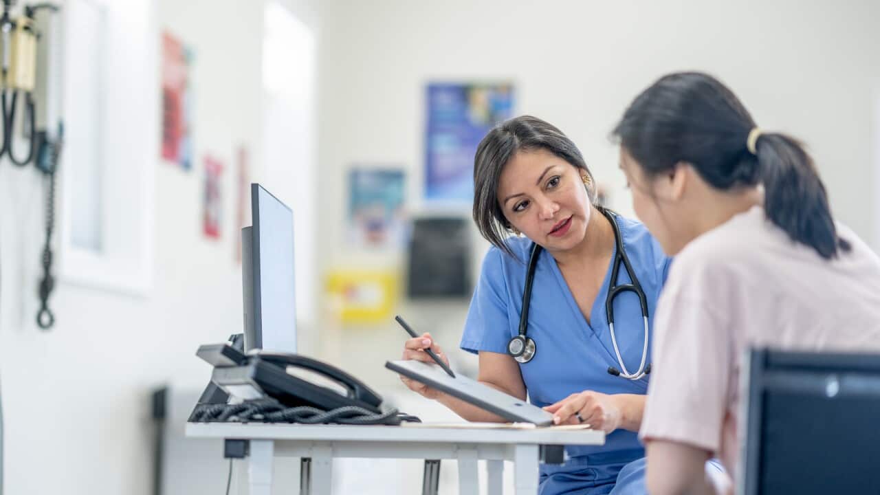 A woman in blue scrubs speaks to a woman seated who has her back to the camera