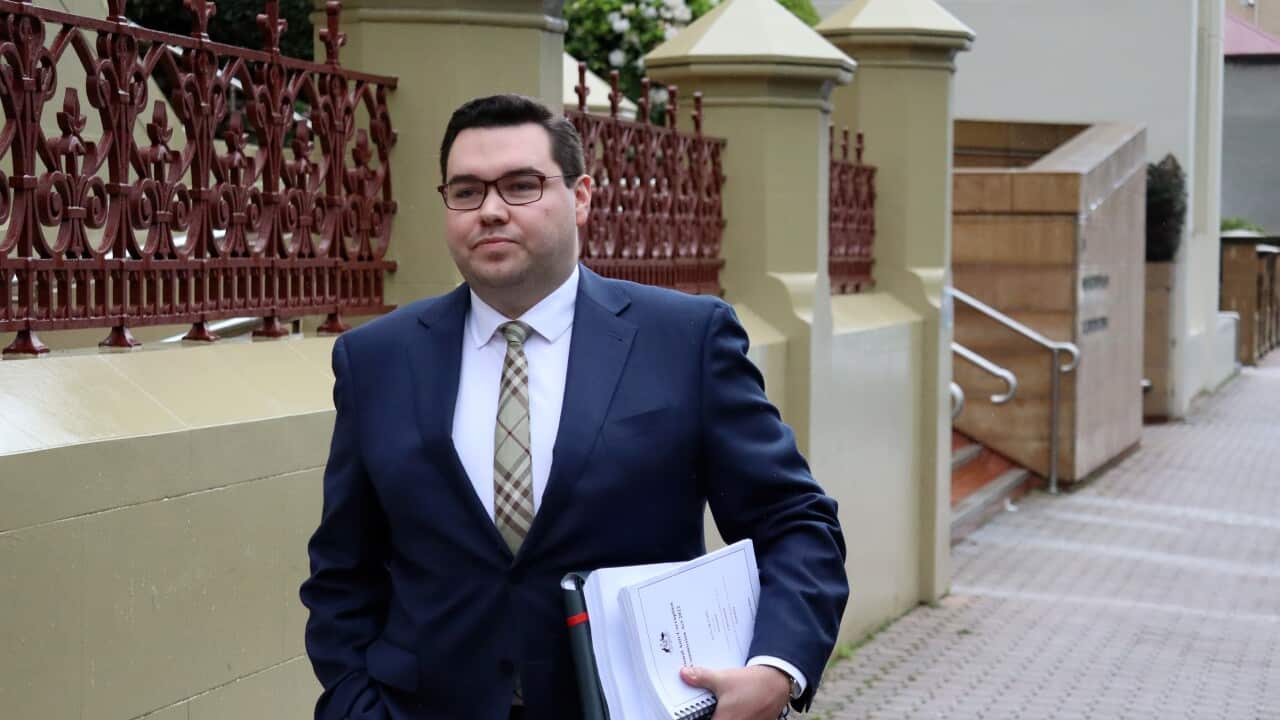 A young white man in navy suit and wearing glasses walking on a footpath. He is carrying a golders and note book.