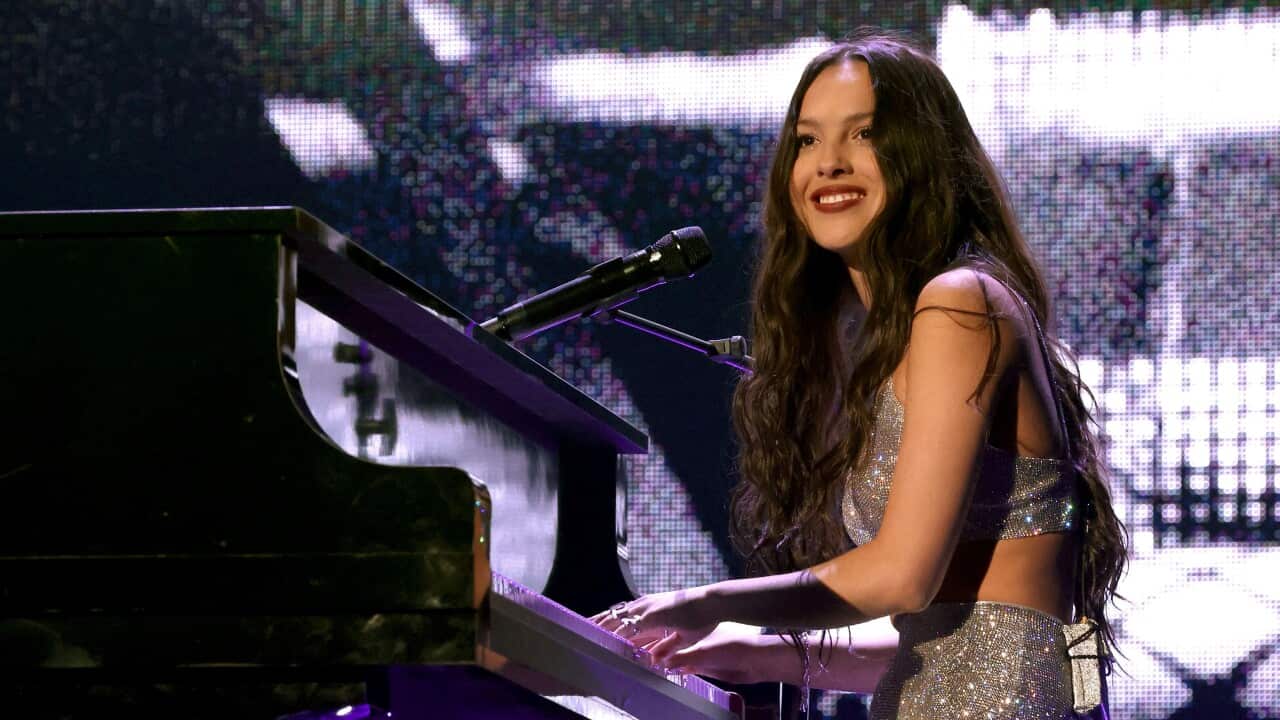 A brunette woman sits at a piano smiling with her hands on the keys.