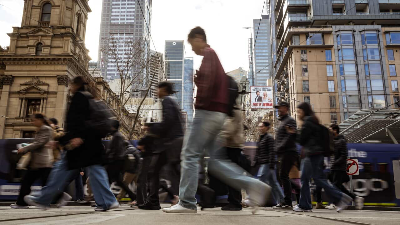 Blurred pedestrians crossing a busy Melbourne city street, with trams, historic buildings and modern skyscrapers in the background.