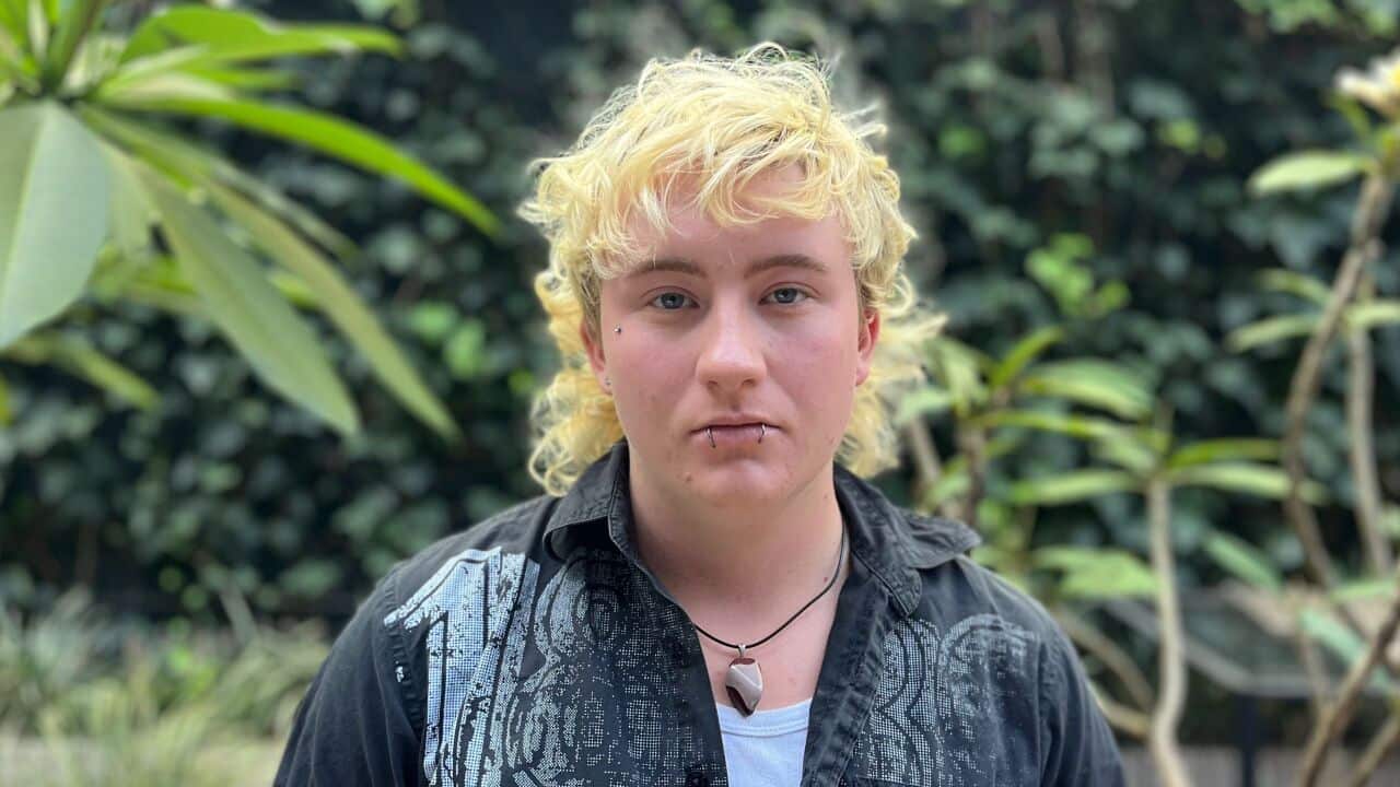 a young man with long blonde hair and lip piercings in front of a green garden