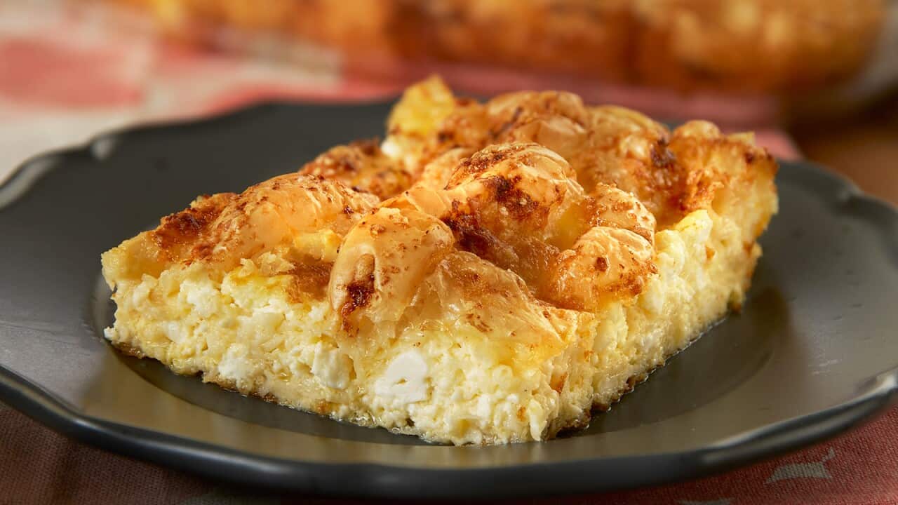 Square piece of pie with soft feta base and slightly scrunched golden-brown filo top sits on dark brown plate.