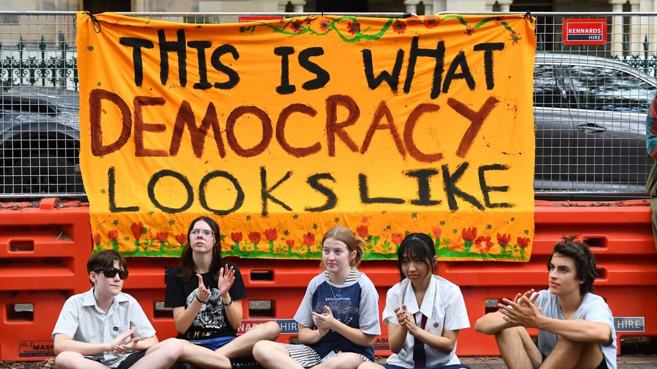 Five teenagers are sitting on the ground under a banner that reads "This is what democracy looks like" next to a construction site.