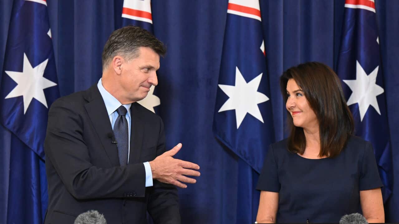 Man wearing a suit extends hand for handshake with woman standing next to him. Both standing in front of Australian flags.