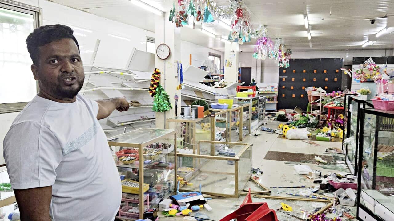 A store owner points behind him at broken shelves.