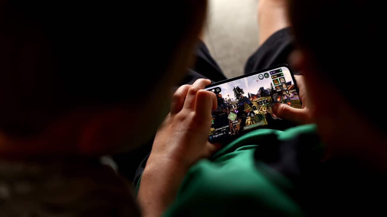 Two children play Roblox on an iPhone. The image is taken over their shoulders, the screen placed between them. One child watches the other play.