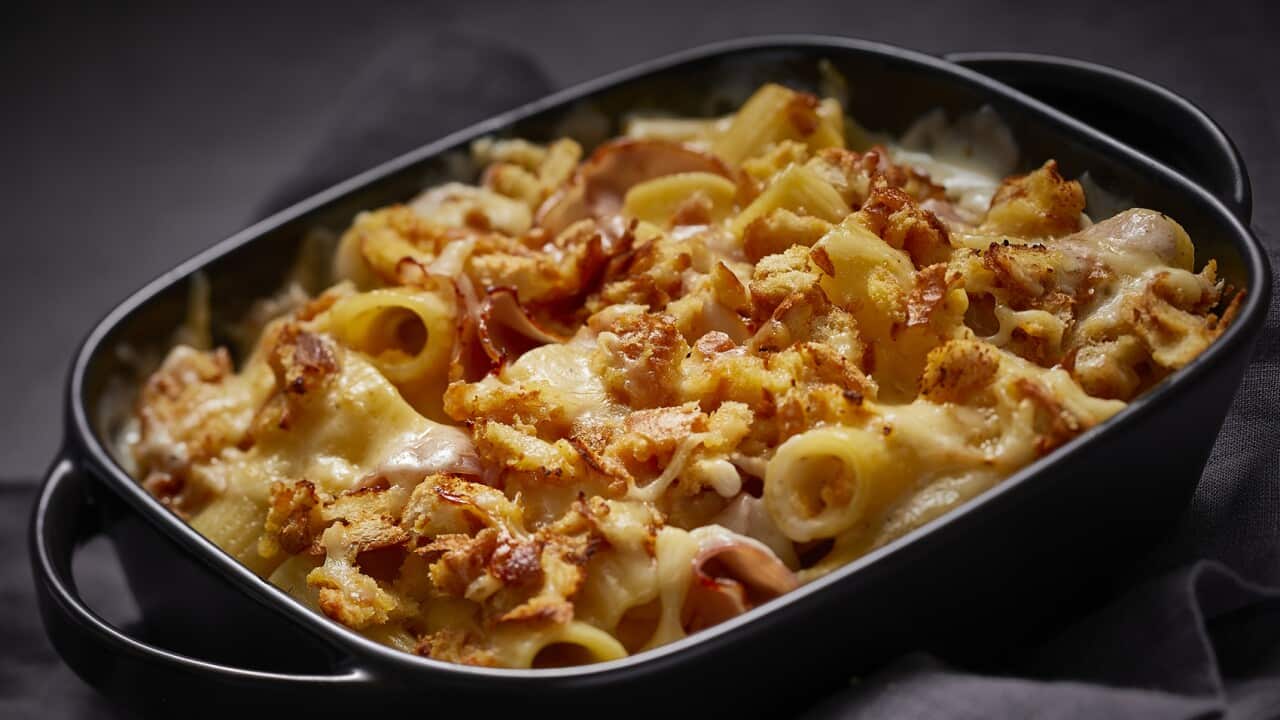 A dark baking dish holds a golden rigatoni pasta bake, with a crunchy cheese and torn bread topping.