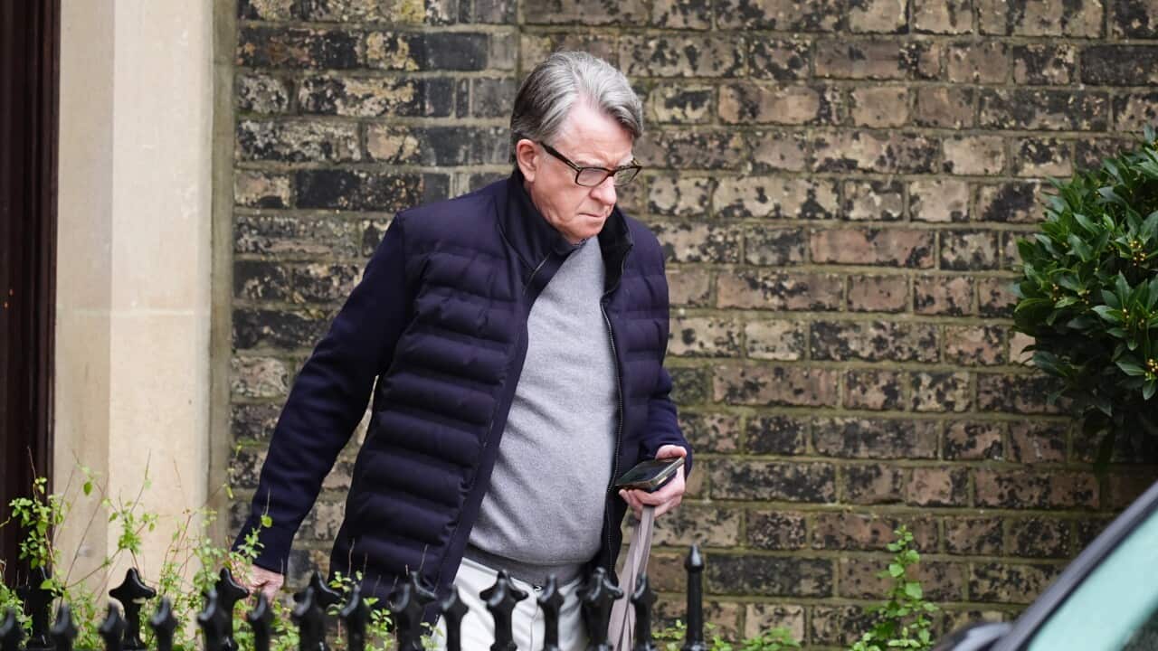 An older man wearing glasses and a dark jacket walks past a brick wall while carrying a folded umbrella and phone.