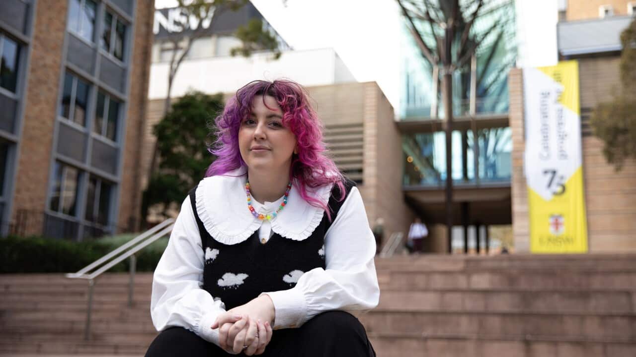 A non-binary person with long pink and purple hair sits on the steps in front of a building.