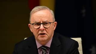 An older man in a dark suit, glasses and striped tie, speaking during a meeting or press event with flags blurred in the background.