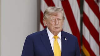 Donald Trump, wearing a white shirt, yellow tie, and blue blazer with an American flag pin on it. He is standing in front of the American flag.