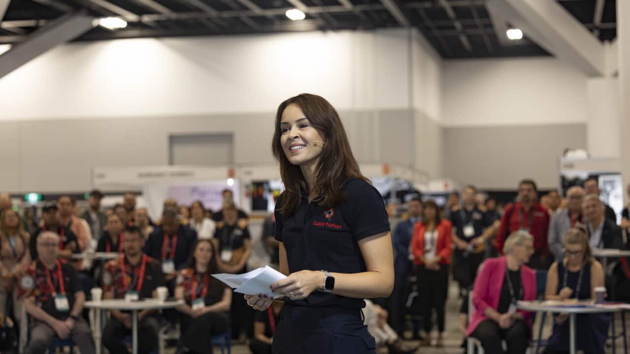 A woman stands before a crowd at an Indigenous business trade show, while holding a piece of paper and preparing to speak.
