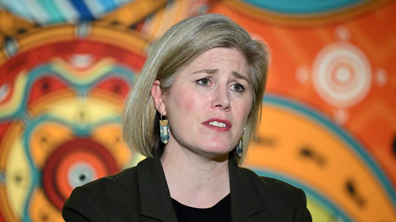 A woman in a black suit with blonde hair stands against a colourful background