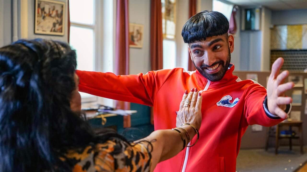 A smiling young man in a bright orange-red tracksuit attempts to embrace his mother, who has a hand out to stop him.
