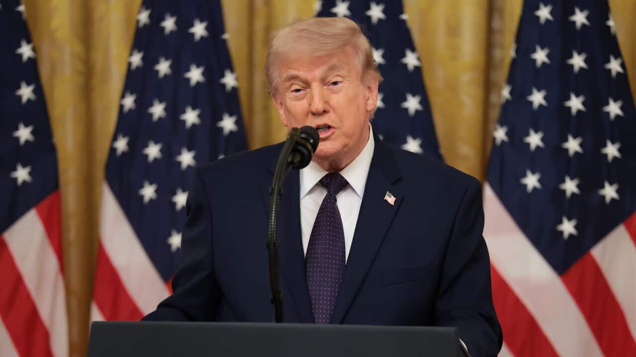 An older white man with light blonde hair wearing a suit and speaking at a podium. US flags hang behind him.