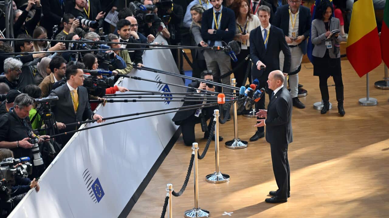 Man in suit stands in front of microphones and crowd of people.
