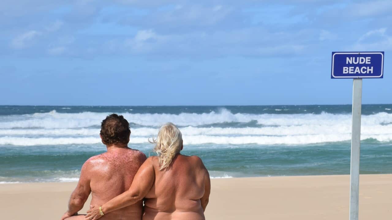 Photo taken from behind of a man and woman on a beach with no shirts on, and a sign saying 'nude beach'.