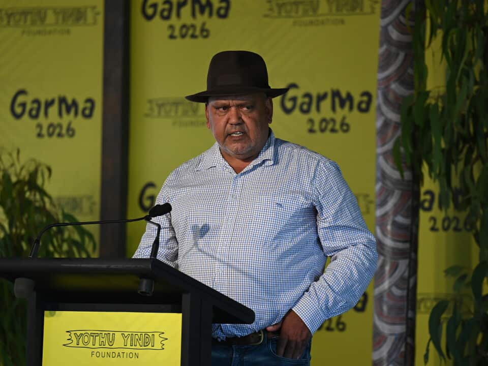 A man wearing a black hat, checked long-sleeve shirt and jeans speaks at a podium during the Garma Festival 2026. Behind him are bright yellow backdrops displaying "Garma 2026" and the Yothu Yindi Foundation logo, with native plants framing the stage. He appears to be addressing an audience with a serious expression.