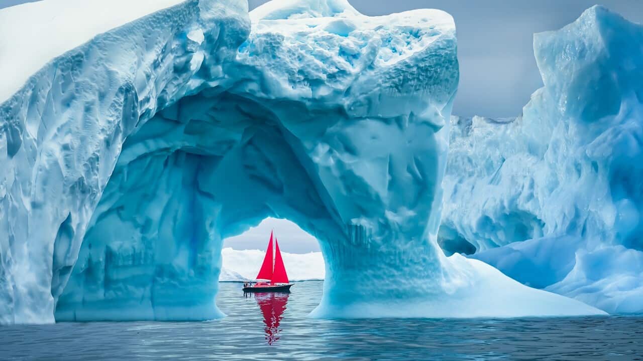 Red sailboat sailing under a majestic iceberg arch on sunny blue Artic Ocean in Greenland, Ilulissat Icefjord, Ilulissat, Disko Bay, Unesco World Heritage Site
