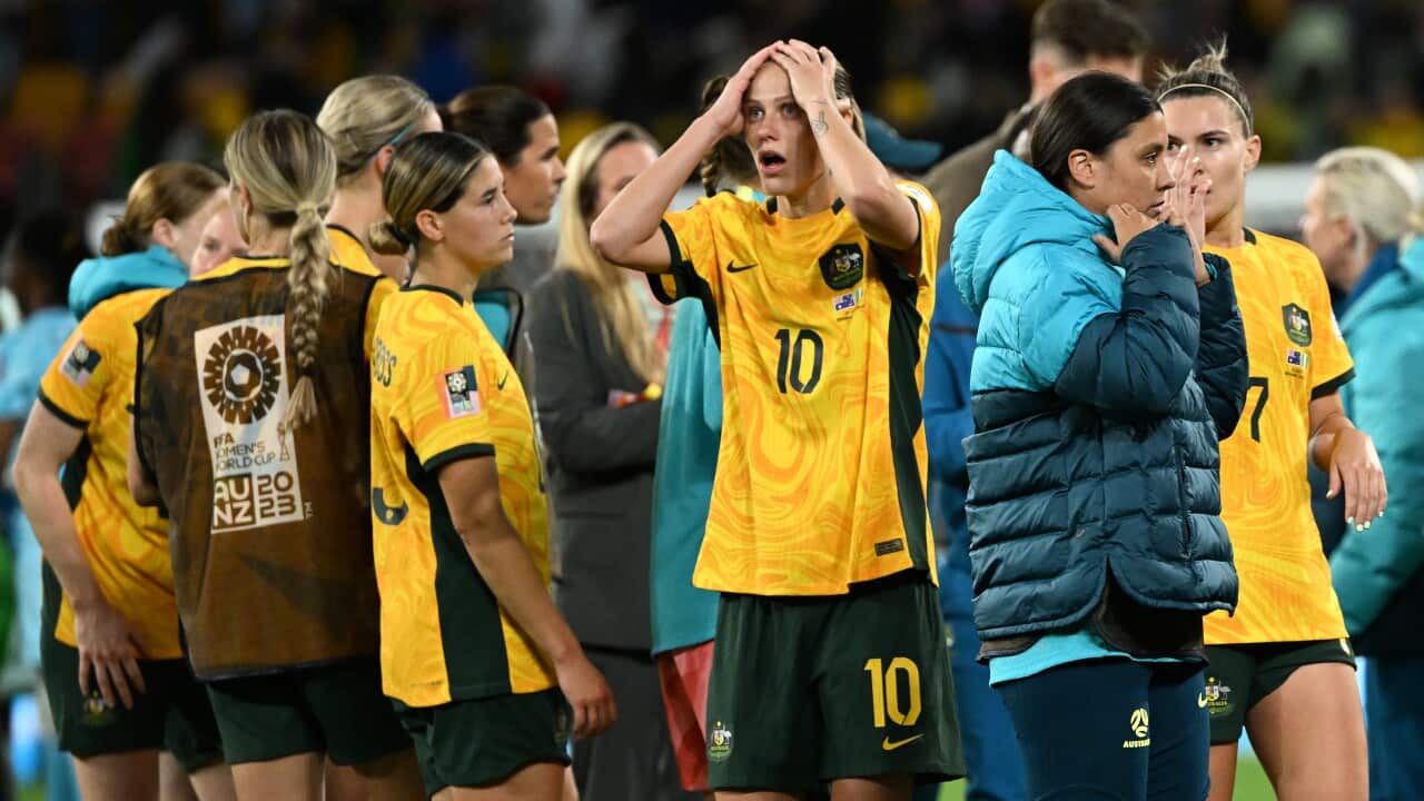 Group of Matildas players huddle together. Woman in middle of group holds her hands to the top of her face, with a shocked expression.