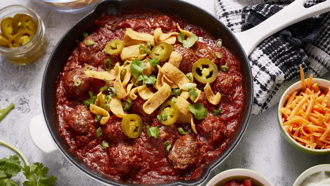 A white-handle cast iron skillet holds meatballs in a rich tomato sauce, topped with corn chip pieces, sliced chilli and coriander leaves. Small bowls with other toppings, including cheese and sour cream, sit alongside.