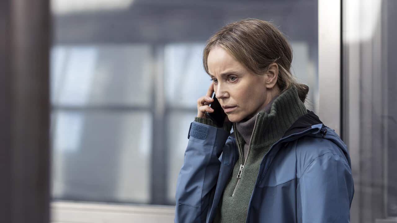 A woman in a heavy blue jacket stands in front of a grey-toned window, talking on a mobile phone.