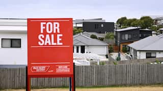A red "For Sale" sign stands in the foreground of a suburban neighbourhood, with modern houses, fenced backyards and an overcast sky in the background.