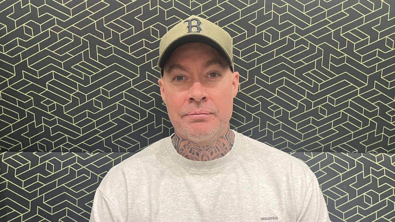 A middle aged man in a grey sweat shirt, neck tattoos and green cap sits in front of printed background