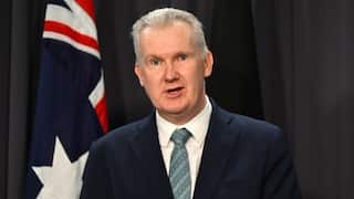 A man in a suit standing in front of an Australian flag and dark blue curtain.