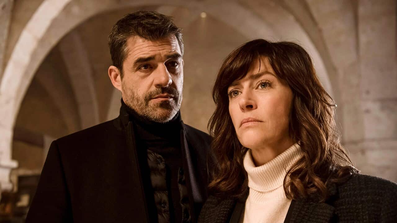 A man and a woman stand in what looks like a church or a historic building, with a stone arch behind them.