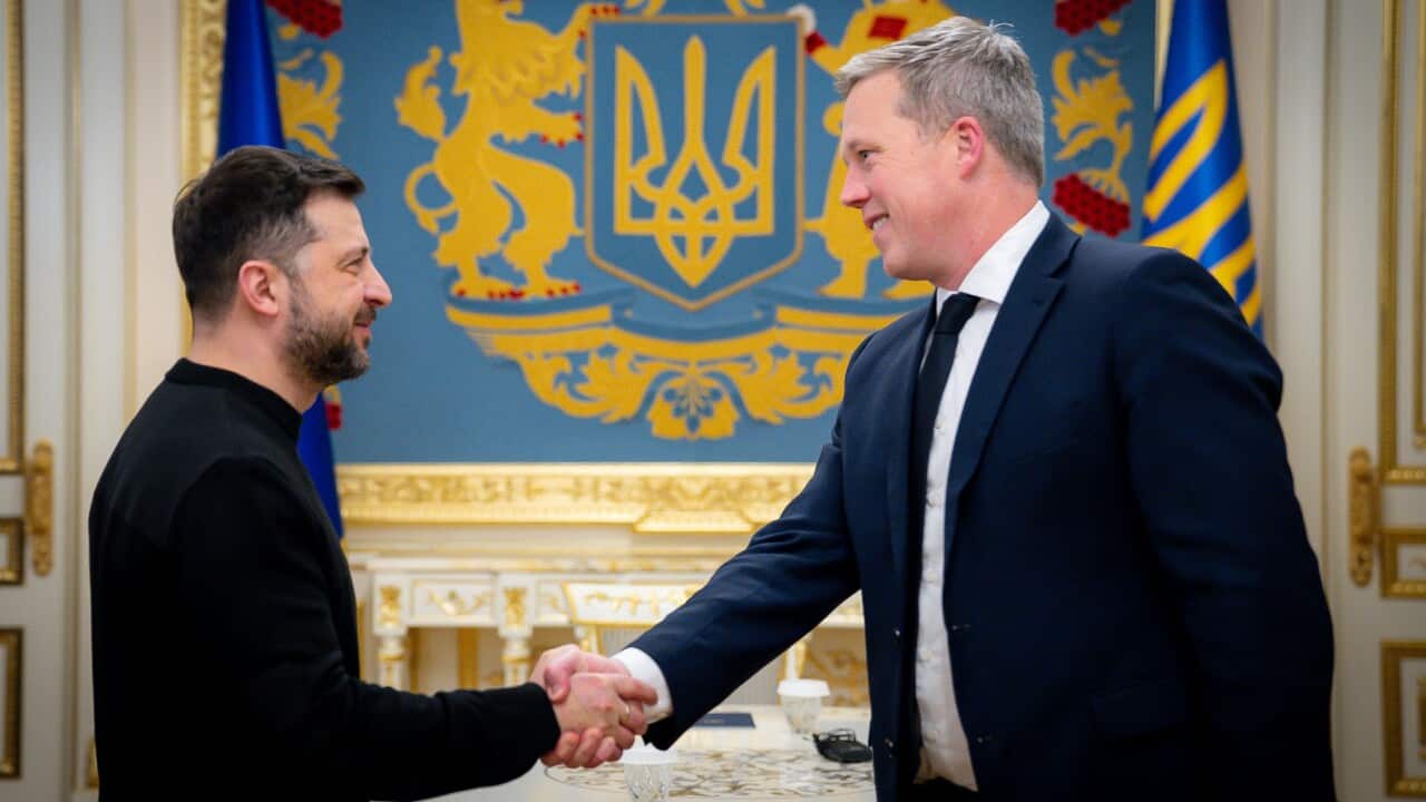 Ukrainian President Volodymyr Zelenskyy, wearing a black shirt, shakes hands with a US official wearing a dark suit and tie, in front of a blue wall featuring the Coat of Arms of Ukraine.