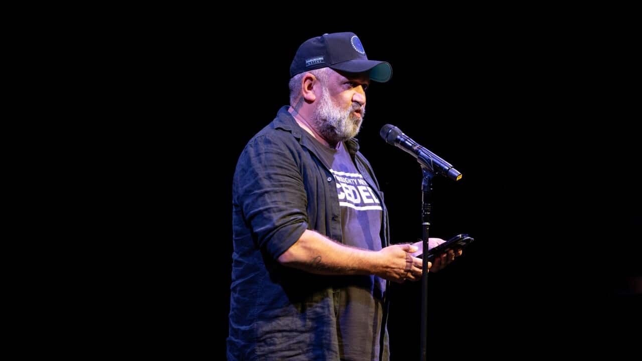 A middle-aged man dressed in black reads from his phone into a microphone