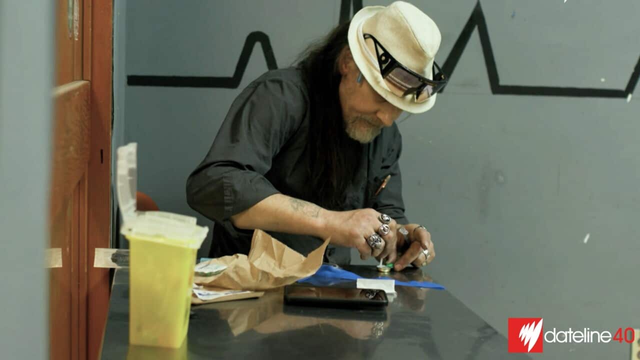 A man sitting at a steel bench preparing something with his hands. There is a yellow container on the bench in the foreground.