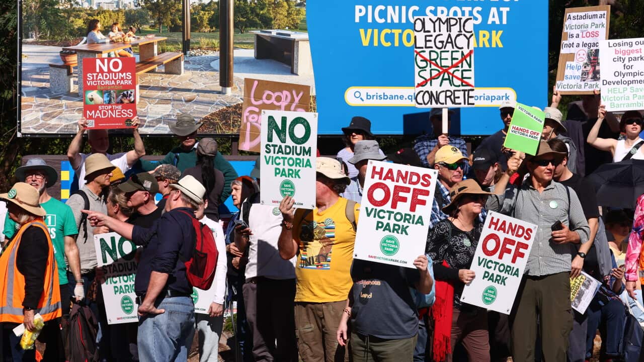 Protesters holding signs opposing a new Olympic stadium at Victoria Park gather in front of a billboard promoting the park.