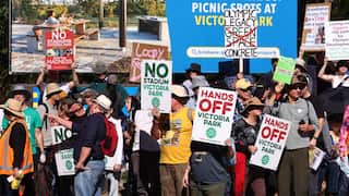 Protesters holding signs opposing a new Olympic stadium at Victoria Park gather in front of a billboard promoting the park.
