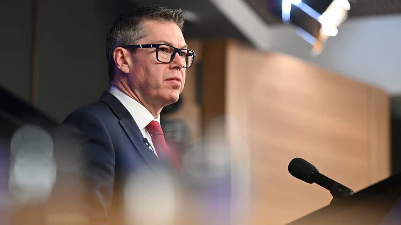 A man with glasses and a red tie stands at a podium speaking into a microphone.