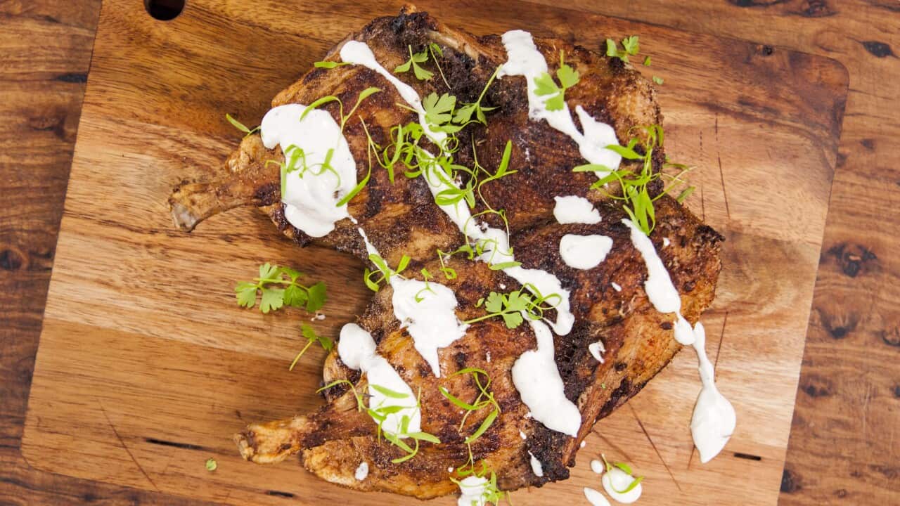 A cooked butterflied duck sits on a wooden board, drizzled with a white sauce and scattered with herb leaves.