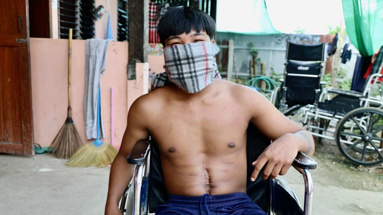 A topless man sitting in a wheelchair, with a grey bandana covering his face below the eyes and a scar running down his stomach.
