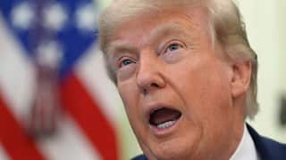 Close-up of an older man with light blond hair speaking mid-sentence, mouth open, with a blurred American flag in the background.