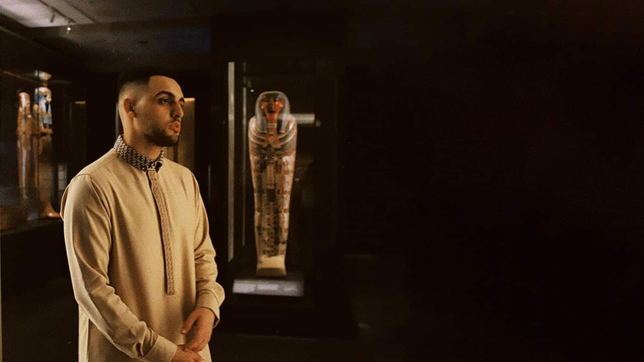 Man in long robe standing in front of Egyptian artefacts.