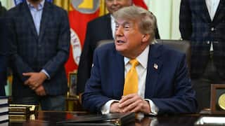 Donald Trump sitting at his desk at the Oval Office wearing a yellow tie surrounded by men in suits.