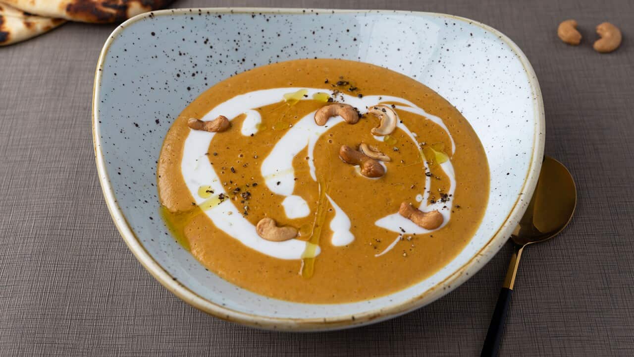 A lovely roundish pottery bowl holds a brown soup drizzled wiht cream and a little golden oil, plus a few cashews. Toasted naan can be seen behind.