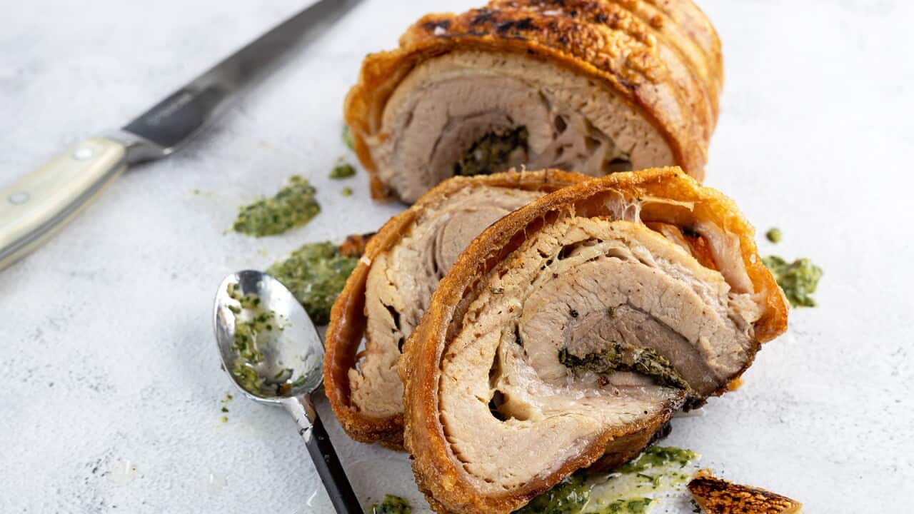 Roast pork with golden crackling sits on a marble surface, several cut slices resting in the front. A spoon sits beside the slices, as if it has just been used to add a few puddles of the green sauce that sit beside the pork.