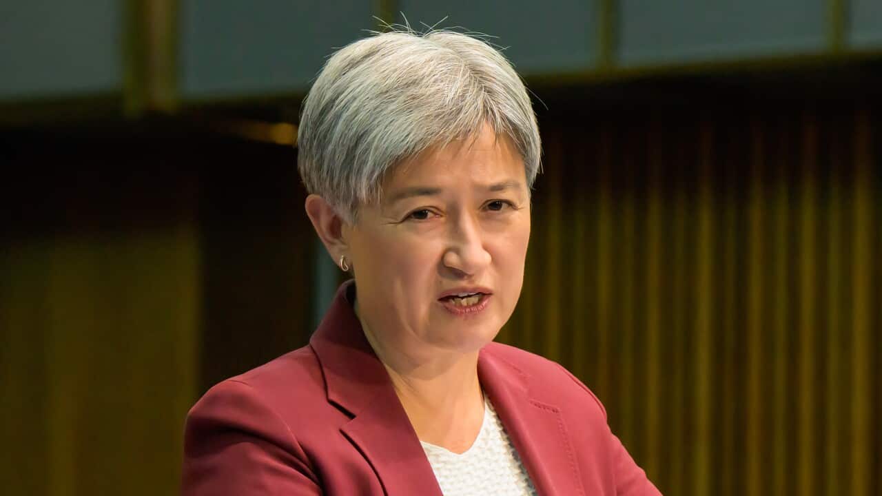 Penny Wong speaking at a lectern in a wood-panelled room wearing a red suit and a white top