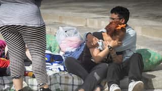 A woman and a man sit on the street, she holds her face and leans on his chest as he comforts her.