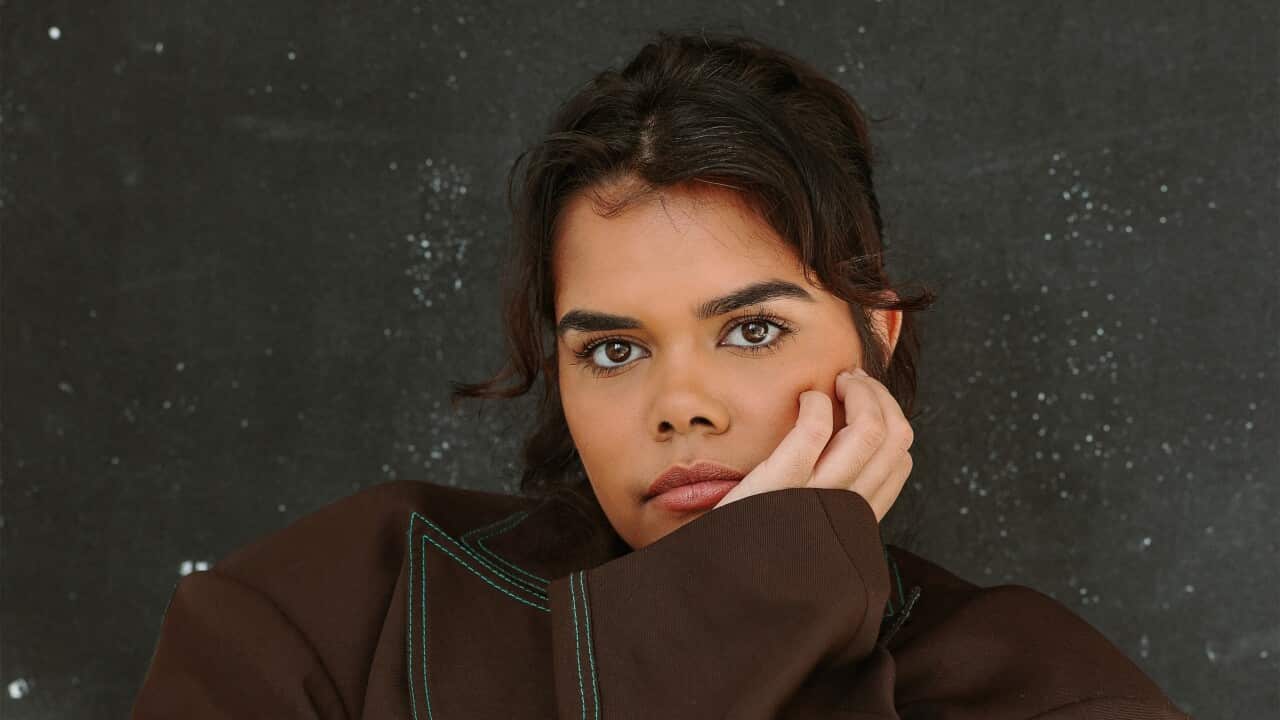 A woman wearing a brown jacket with green seams sits her head in one of her hands with the other arm crossed across her body.