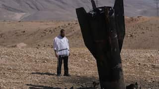 A person stands beside the remains of a large projectile embedded in the ground in a dry, mountainous area.