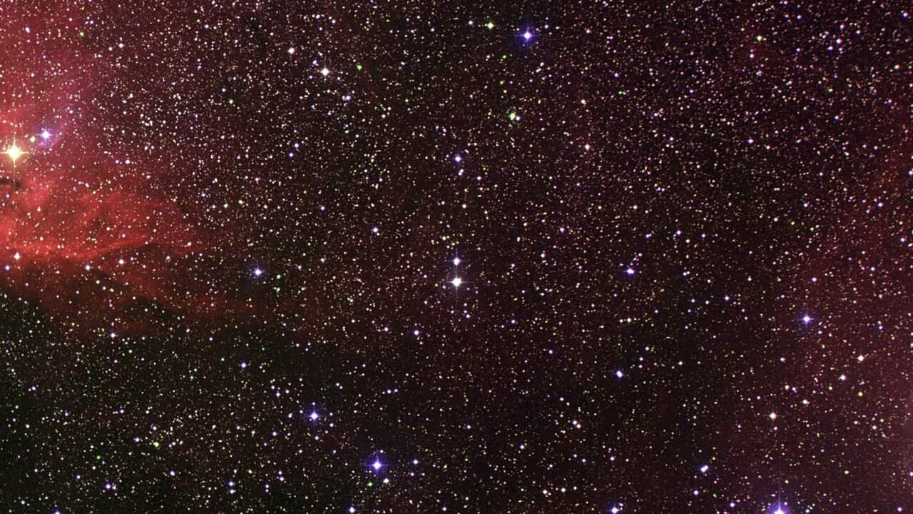 A deep field photograph of space. The image is filled with stars and galaxies shining bright white against a maroon background. To the left of the image is a bright red nebula with a yellow star at its centre.