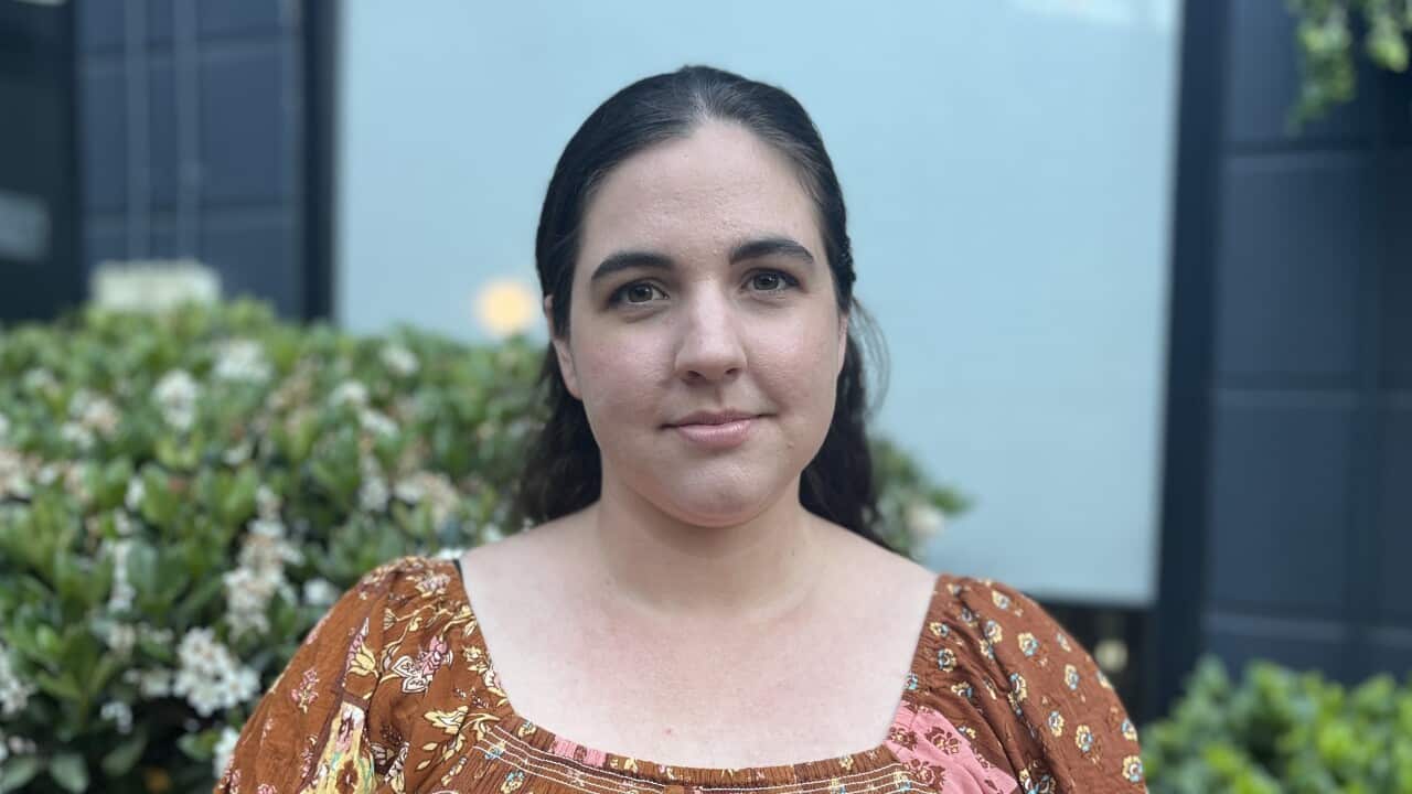 a young woman in a printed dress and brown hair tied back stands in a garden and subtly smiles at the camera